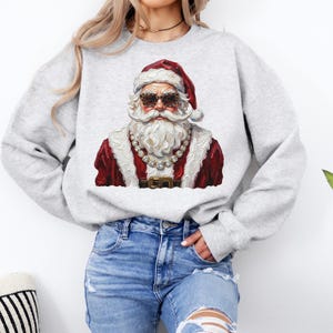 May include: A light gray crewneck sweatshirt featuring a graphic print of Santa Claus wearing leopard print sunglasses and a large crystal-style chain necklace. The design depicts Santa in a red coat with white fur trim and a matching red hat.