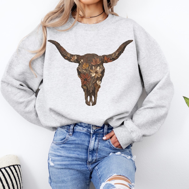 May include: A light gray sweatshirt featuring a detailed illustration of a bull skull adorned with floral accents in shades of brown, orange, and yellow. The skull design is centered on the front of the long-sleeved top.