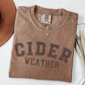 K&ouml;nnte beinhalten: Ein braunes Comfort Colors T-Shirt mit den Worten "CIDER WEATHER" in dunkelbraunen Blockbuchstaben aufgedruckt. Eine silberne Halskette mit einem kleinen Anh&auml;nger liegt &uuml;ber dem Shirt. Ein brauner Hut ist in der oberen linken Ecke zu sehen.