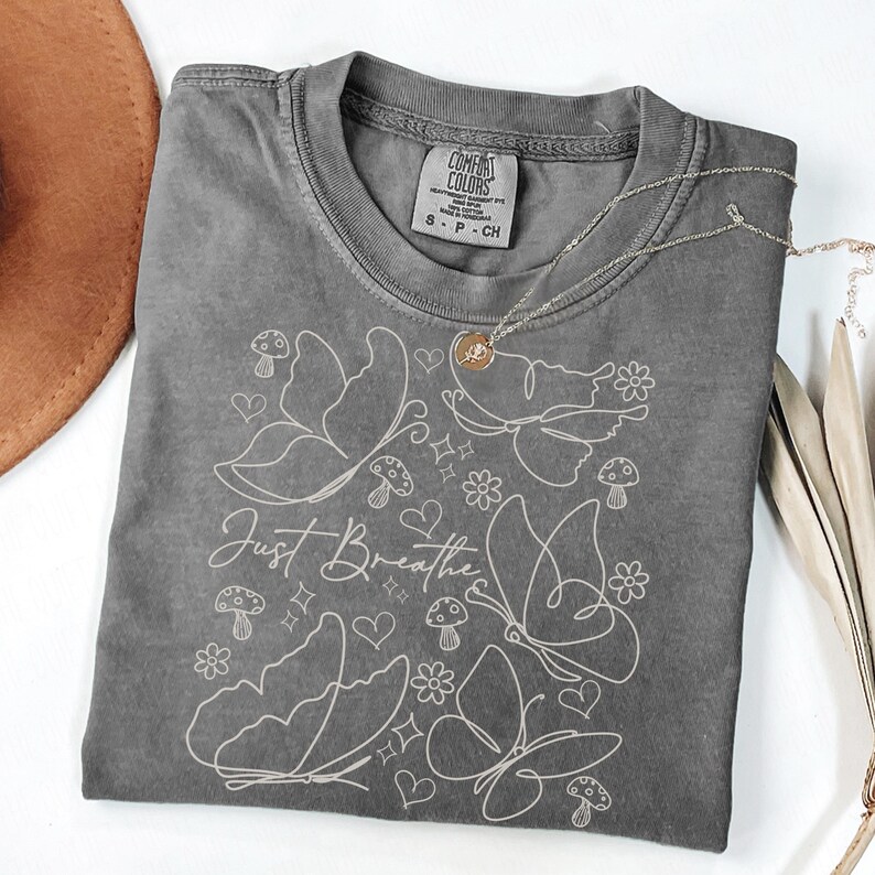 Puede incluir: Una camiseta gris Comfort Colors con un dise&ntilde;o de mariposas y flores blancas. La camiseta presenta las palabras "Just Breathe" en una fuente cursiva. Un collar de oro con un colgante est&aacute; drapeado sobre la camiseta.