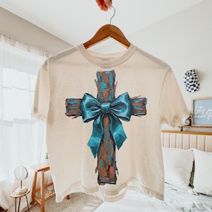 Peut inclure: Un t-shirt cr&egrave;me avec un motif de croix peinte et un n&oelig;ud bleu sarcelle. La croix a un aspect textur&eacute; en bois avec des nuances de marron et d'orange. Le n&oelig;ud bleu sarcelle ajoute une touche de couleur sur le fond neutre du t-shirt.
