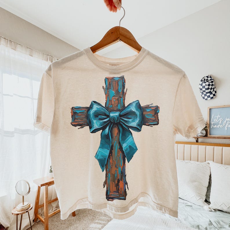Peut inclure: Un t-shirt cr&egrave;me avec un motif de croix peinte et un n&oelig;ud bleu sarcelle. La croix a un aspect textur&eacute; en bois avec des nuances de marron et d'orange. Le n&oelig;ud bleu sarcelle ajoute une touche de couleur sur le fond neutre du t-shirt.