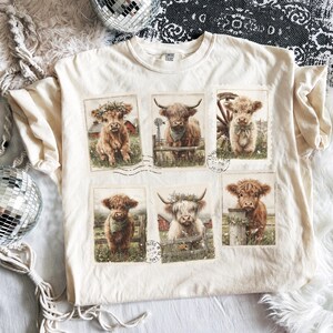 May include: Cream-colored t-shirt featuring six rectangular images of Highland cows. Each image showcases a different cow with floral accents, set against a rural backdrop. The shirt has a classic crew neck and short sleeves.