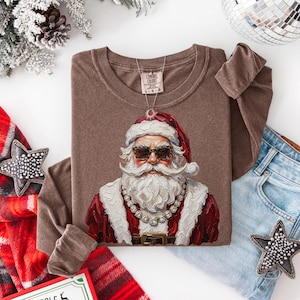 May include: A brown long-sleeve t-shirt featuring a graphic of Santa Claus wearing leopard print sunglasses and a gold chain necklace. The shirt has a crew neckline and a Comfort Colors brand tag at the inner collar. The graphic is painted in style.