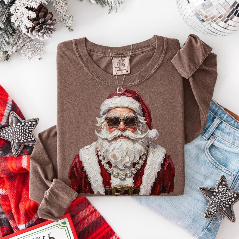 May include: A brown long-sleeve t-shirt featuring a graphic of Santa Claus wearing leopard print sunglasses and a gold chain necklace. The shirt has a crew neckline and a Comfort Colors brand tag at the inner collar. The graphic is painted in style.