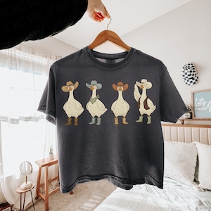 May include: Dark gray t-shirt featuring a cartoon illustration of four geese dressed as cowboys. Each goose wears a cowboy hat, boots, and one has a bandana. The shirt is hanging on a wooden hanger.