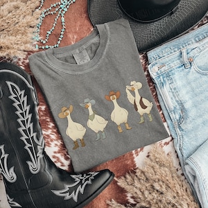 May include: A gray t-shirt featuring four cartoon geese wearing cowboy hats and boots. The shirt is surrounded by a black cowboy boot, a black hat, and blue jeans, creating a Western theme.