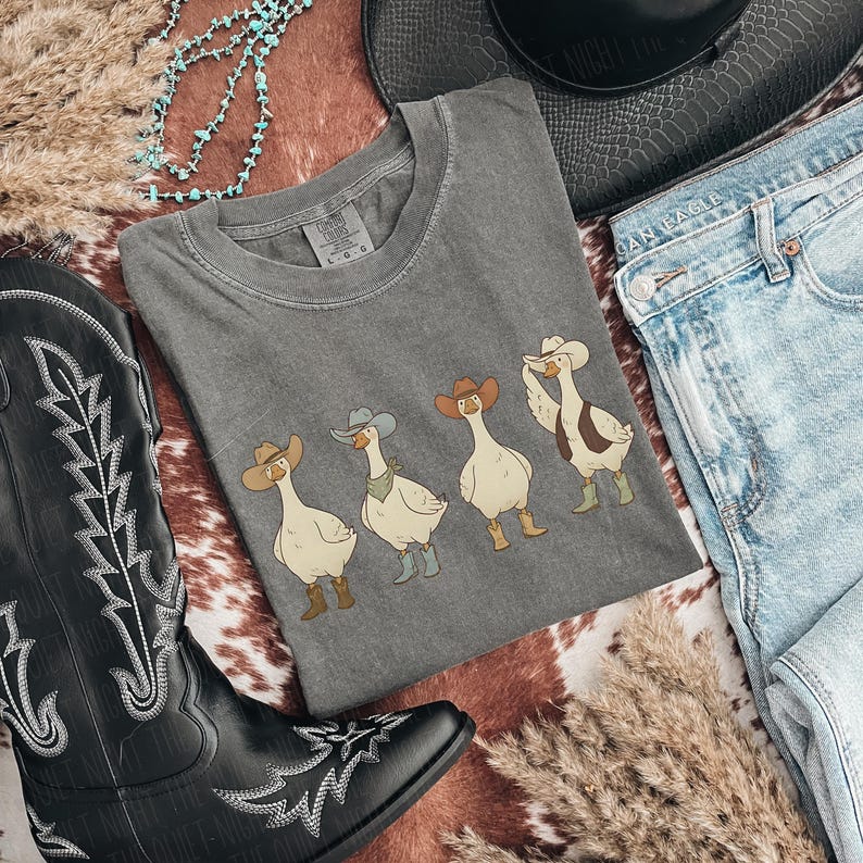 May include: A gray t-shirt featuring four cartoon geese wearing cowboy hats and boots. The shirt is surrounded by a black cowboy boot, a black hat, and blue jeans, creating a Western theme.