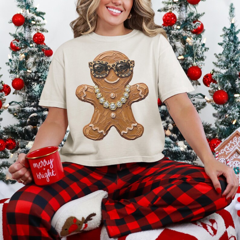 May include: A cream-colored t-shirt featuring a graphic of a gingerbread person wearing leopard print sunglasses and a rhinestone necklace. The gingerbread person has white icing details on its arms and legs. The shirt is worn with red and black plaid pants.