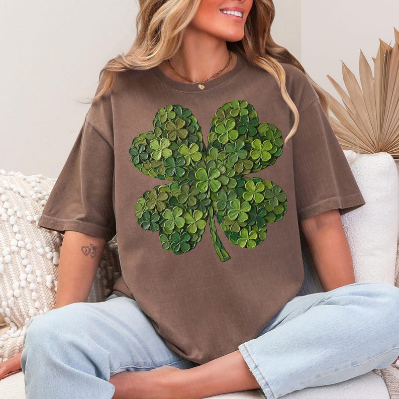 May include: A brown t-shirt featuring a large, detailed four-leaf clover design. The clover is composed of many smaller green clovers, creating a textured, layered effect. The shirt has short sleeves and a relaxed fit, suitable for casual wear.