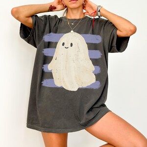 May include: A charcoal gray short-sleeve t-shirt featuring a central graphic of a cream-colored ghost with a simple smiling face. The ghost is positioned over four horizontal light purple brushstroke stripes that span the width of the chest.