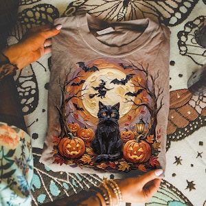 May include: A tan t-shirt featuring a graphic print of a black cat sitting among orange jack-o'-lanterns, bare trees, and fallen leaves. A large full moon glows in the background with a silhouette of a witch on a broomstick and flying bats.