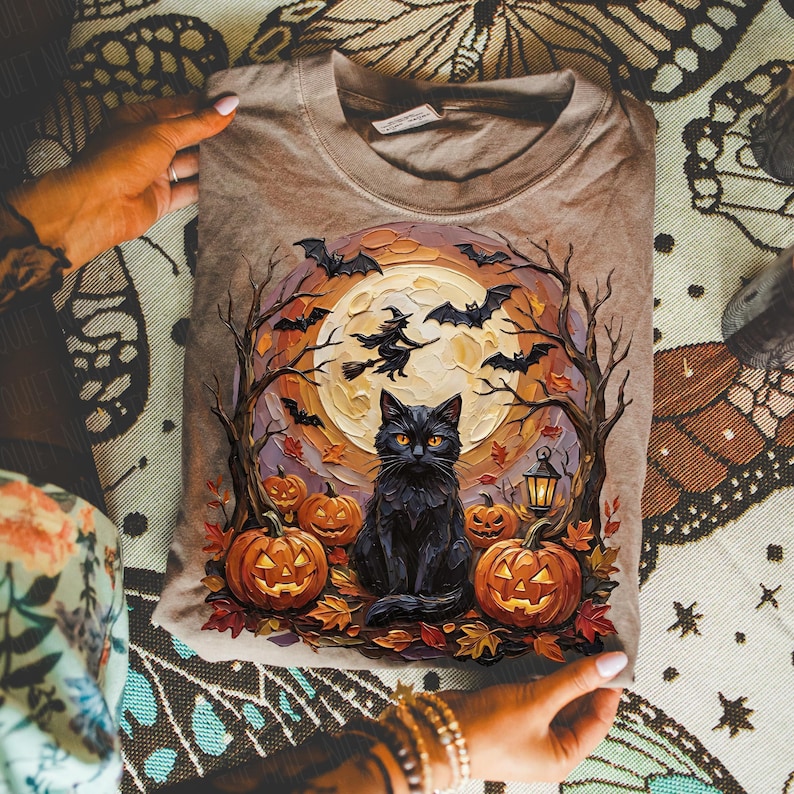 May include: A tan t-shirt featuring a graphic print of a black cat sitting among orange jack-o'-lanterns, bare trees, and fallen leaves. A large full moon glows in the background with a silhouette of a witch on a broomstick and flying bats.