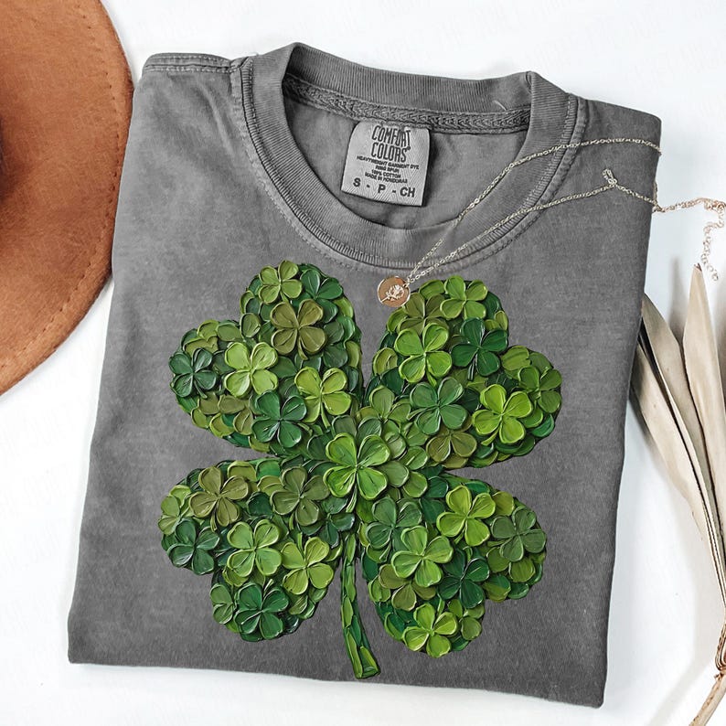 May include: A gray Comfort Colors t-shirt featuring a large, detailed four-leaf clover design in various shades of green. A delicate silver necklace with a small pendant rests on the shirt. The shirt is size S-P-CH.