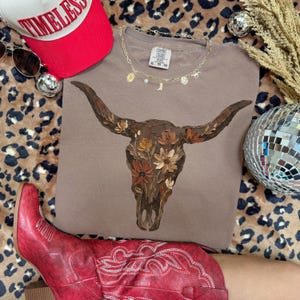 May include: A taupe Comfort Colors t-shirt featuring a detailed illustration of a bull skull adorned with flowers in shades of brown, orange, and cream. The shirt is styled with a red baseball cap, sunglasses, and a gold necklace.