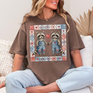 Pu&ograve; includere: T-shirt marrone con un'illustrazione ad acquerello di due procioni che si tengono per mano e un palloncino rosso a forma di cuore. I procioni indossano salopette blu e sono incorniciati da un bordo patchwork con cuori.