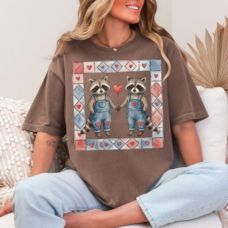 Pu&ograve; includere: T-shirt marrone con un'illustrazione ad acquerello di due procioni che si tengono per mano e un palloncino rosso a forma di cuore. I procioni indossano salopette blu e sono incorniciati da un bordo patchwork con cuori.