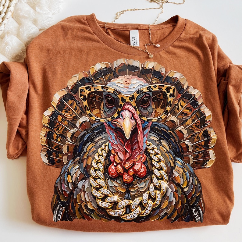 May include: A rust-colored t-shirt featuring a graphic print of a turkey wearing leopard-print glasses and a thick, sparkling gold chain. The turkey has detailed, textured feathers in shades of brown, black, and tan, with a bright red wattle.