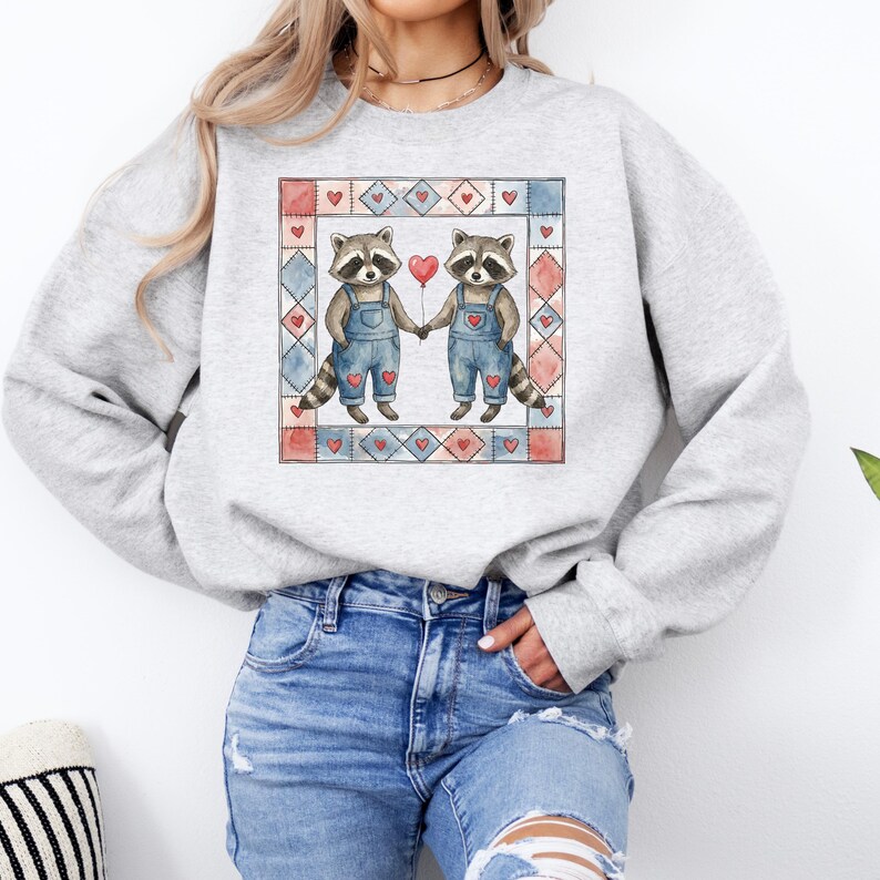 Pu&ograve; includere: Una felpa grigio chiaro con un'illustrazione ad acquerello di due procioni che si tengono per mano e un palloncino rosso a forma di cuore. I procioni indossano salopette blu, su un design patchwork con cuori.