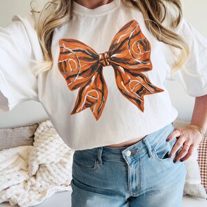 May include: White t-shirt featuring a large orange and black bow design. The bow has a textured appearance with a basketball-like pattern. The shirt is paired with light blue jeans.