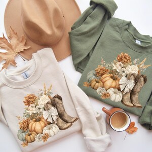 May include: Two sweatshirts, one cream and one olive green, feature a fall-themed design. The design includes pumpkins, flowers, and cowboy boots. A tan hat and a fall leaf are also visible.