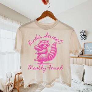 May include: Cream-colored t-shirt with a pink raccoon graphic and the text "Kinda Sweet, Mostly Feral." The raccoon is standing with its arms crossed. The shirt is hanging on a wooden hanger.
