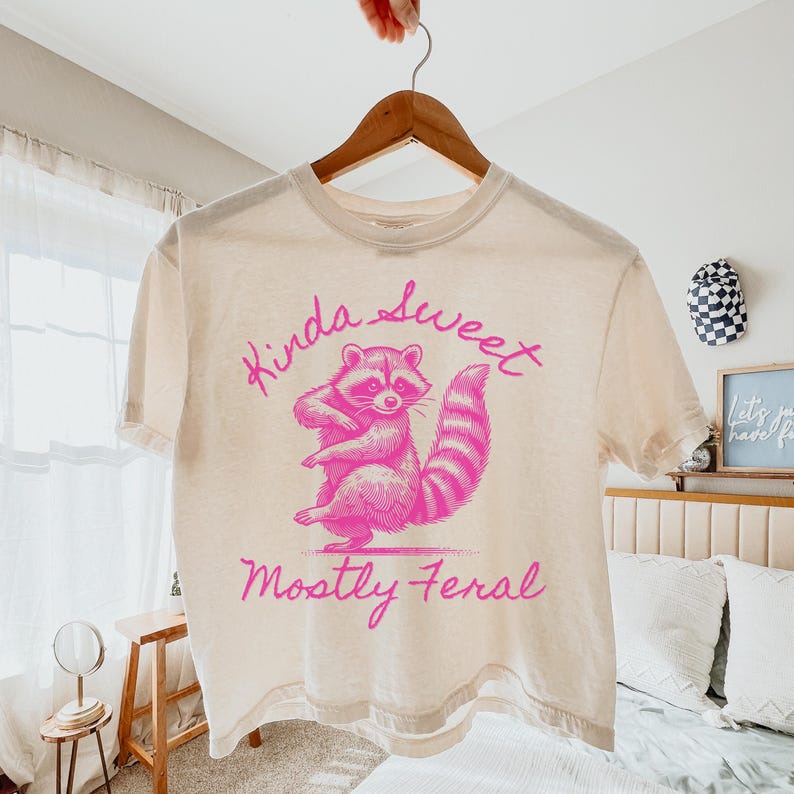 May include: Cream-colored t-shirt with a pink raccoon graphic and the text "Kinda Sweet, Mostly Feral." The raccoon is standing with its arms crossed. The shirt is hanging on a wooden hanger.