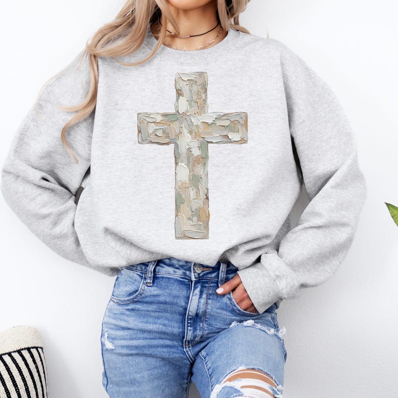 May include: A light gray sweatshirt featuring a large, textured cross design in shades of beige, gray, and green. The cross is the focal point, set against the plain background of the sweatshirt.