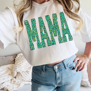 K&ouml;nnte beinhalten: Ein wei&szlig;es Kurzarm-T-Shirt mit dem Wort MAMA in gro&szlig;en Blockbuchstaben. Die Buchstaben haben ein leuchtend gr&uuml;nes und gelbes Leopardenmuster. Das T-Shirt wird mit einer hellblauen Jeans mit einem sichtbaren Metallknopf kombiniert.