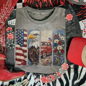 Puede incluir: Camiseta gris de cuello redondo con un dise&ntilde;o que presenta la bandera estadounidense, un &aacute;guila calva, una camioneta roja y un ramo de flores. La camiseta est&aacute; rodeada de vaqueros, un sombrero de vaquero rojo, botas rojas, fichas de p&oacute;quer y dados.