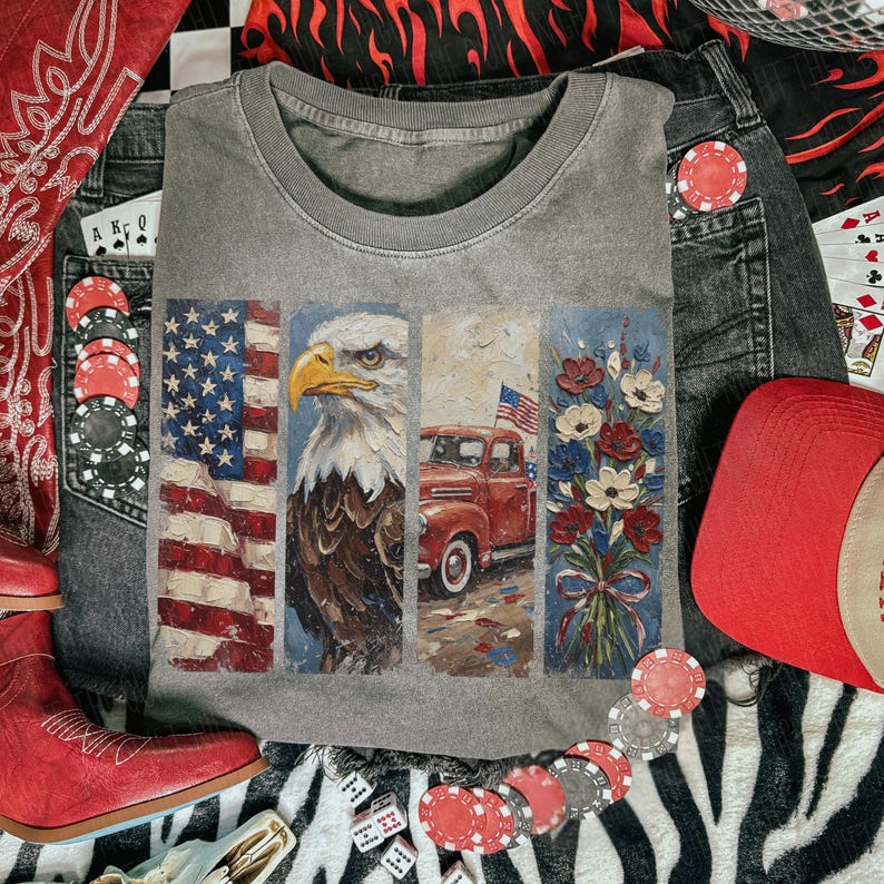 Puede incluir: Camiseta gris de cuello redondo con un dise&ntilde;o que presenta la bandera estadounidense, un &aacute;guila calva, una camioneta roja y un ramo de flores. La camiseta est&aacute; rodeada de vaqueros, un sombrero de vaquero rojo, botas rojas, fichas de p&oacute;quer y dados.