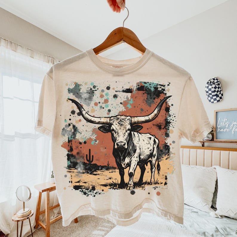 May include: Cream-colored crop top featuring a graphic of a longhorn steer with large horns, set against a backdrop of desert scenery in shades of red, teal, and black. The shirt is hanging on a wooden hanger.