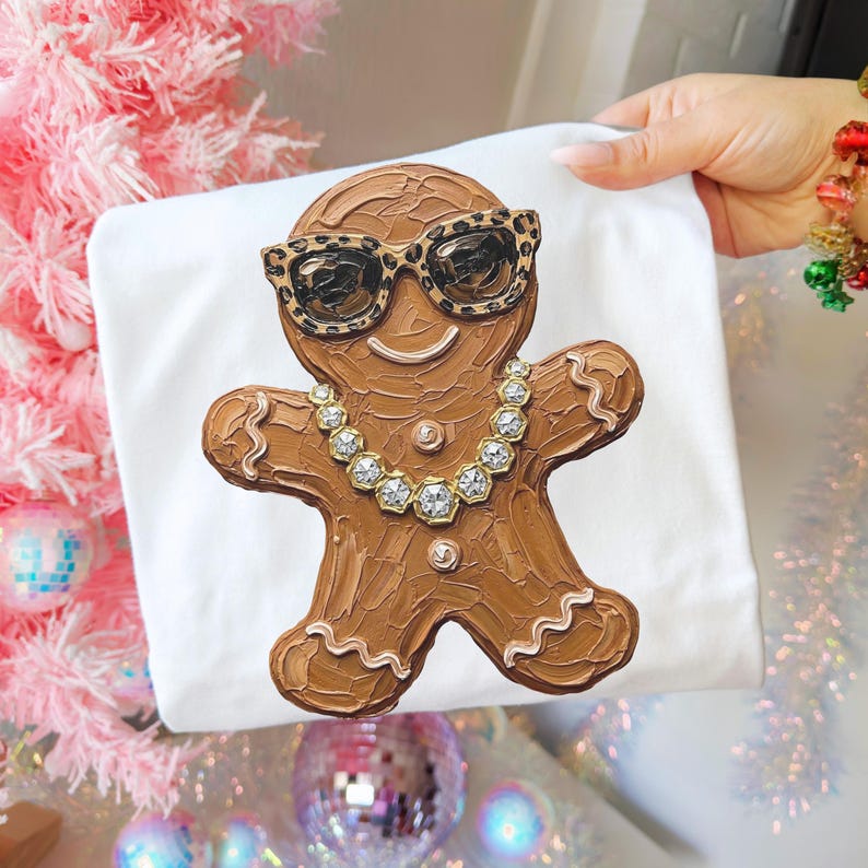May include: A white t-shirt featuring a textured brown gingerbread person graphic. The gingerbread figure wears leopard print sunglasses and a silver-toned chain necklace with clear faceted stones. The design has visible brushstroke textures.