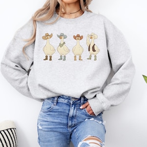 May include: A light gray sweatshirt featuring four cartoon geese dressed as cowboys. Each goose wears a cowboy hat, boots, and a vest or bandana. The geese are in a row, with one goose raising its wing.