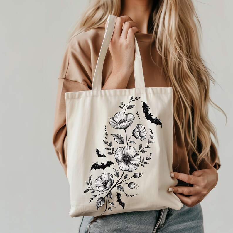 May include: Cream-colored tote bag featuring a black floral design with bat silhouettes. The design includes detailed flowers, leaves, and several bats. The bag has long handles and is held by a person.