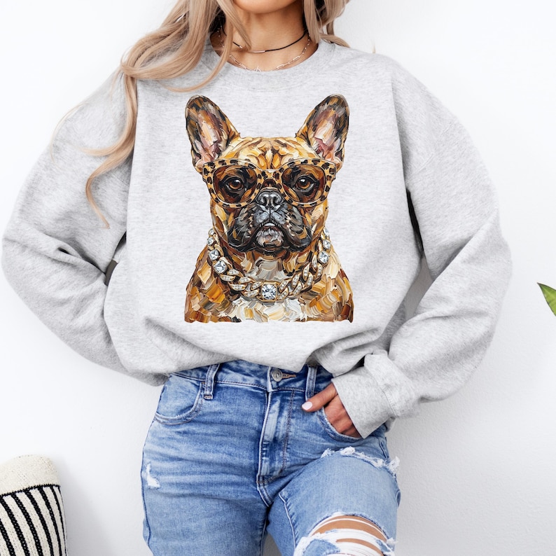 May include: A light gray sweatshirt featuring a detailed illustration of a French Bulldog wearing leopard print glasses and a diamond chain. The dog's fur is a mix of brown and tan tones. The sweatshirt is a casual style.
