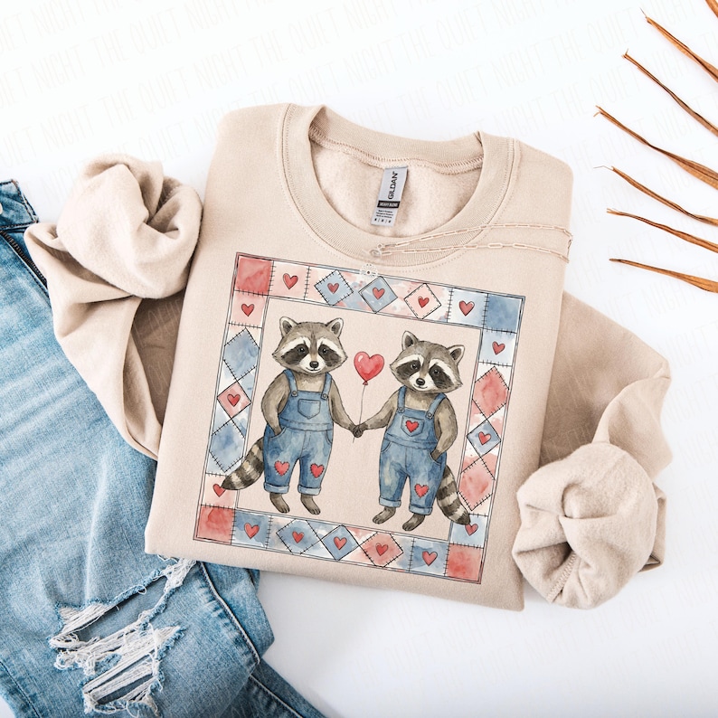 Pu&ograve; includere: Felpa beige con un'illustrazione ad acquerello di due procioni che si tengono per mano e un palloncino rosso a forma di cuore. I procioni indossano salopette blu con dettagli a cuore, all'interno di una cornice patchwork di quadrati rossi e blu.