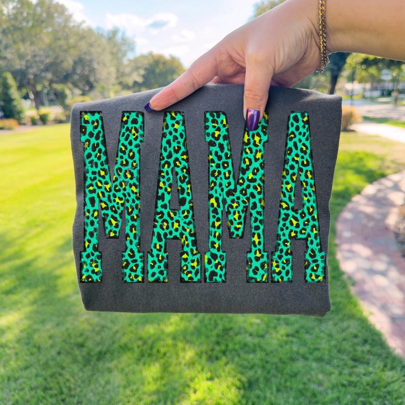 K&ouml;nnte beinhalten: Ein dunkelgraues T-Shirt mit dem Wort MAMA in gro&szlig;en Blockbuchstaben. Die Buchstaben haben ein leuchtendes neongr&uuml;nes und schwarzes Leopardenmuster. Das Design ist auf der Vorderseite des Kleidungsst&uuml;cks zentriert, das vor einem Au&szlig;enbereich gehalten wird.
