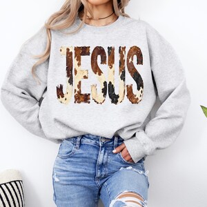 K&ouml;nnte beinhalten: Hellgraues Sweatshirt mit dem Wort "JESUS" im Kuhfellmuster auf der Brust. Das lang&auml;rmlige Oberteil wird mit einer blauen Jeans kombiniert. Das Sweatshirt ist ein l&auml;ssiger, bequemer Stil.