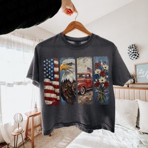 Puede incluir: Camiseta gris oscuro con un dise&ntilde;o patri&oacute;tico. El dise&ntilde;o presenta la bandera estadounidense, un &aacute;guila calva, una camioneta roja con una bandera y un ramo de flores. La camiseta est&aacute; colgada en una percha de madera.