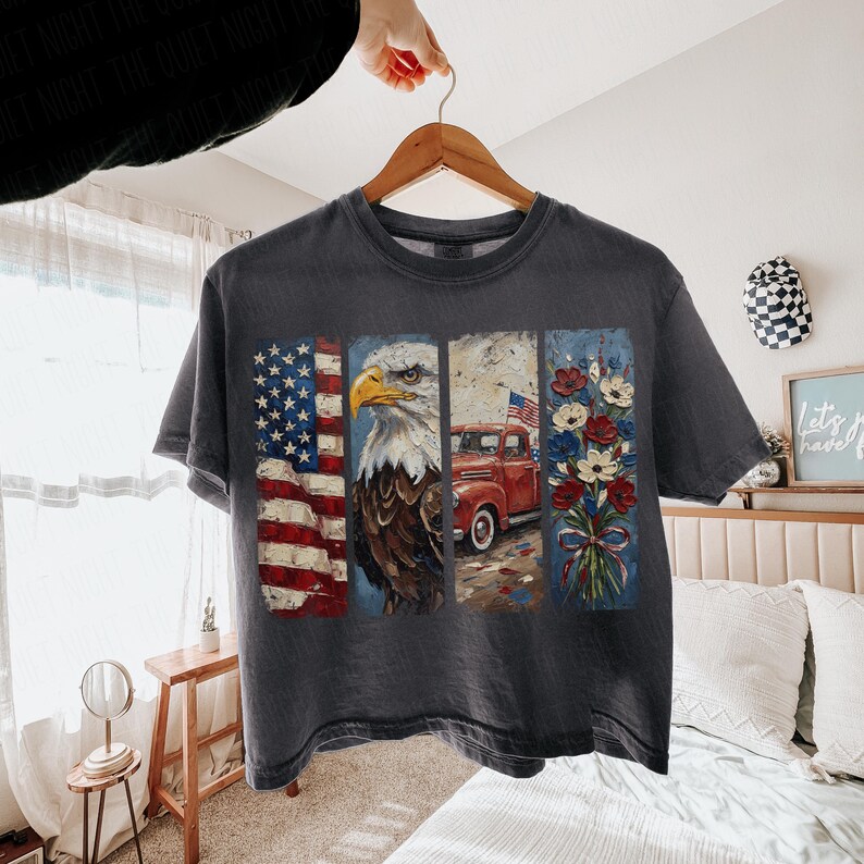 Puede incluir: Camiseta gris oscuro con un dise&ntilde;o patri&oacute;tico. El dise&ntilde;o presenta la bandera estadounidense, un &aacute;guila calva, una camioneta roja con una bandera y un ramo de flores. La camiseta est&aacute; colgada en una percha de madera.