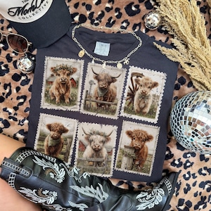May include: Dark gray t-shirt featuring six postage stamp-style images of Highland cows. The shirt is accessorized with a gold necklace, sunglasses, and a black baseball cap. A pair of black cowboy boots with white embroidery are also visible.
