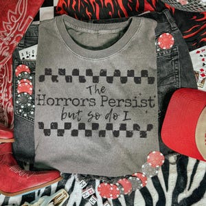 May include: A gray crew neck t-shirt with the text "The Horrors Persist but so do I" in black. The shirt is surrounded by denim shorts, a red trucker hat, red cowboy boots, playing cards, poker chips, and dice.