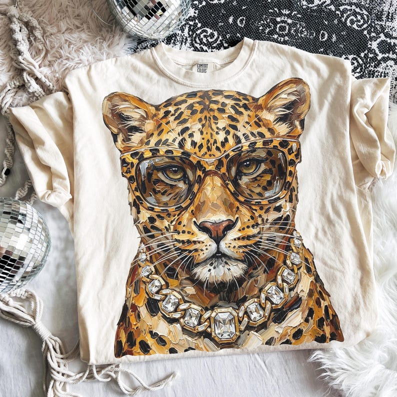 Puede incluir: Camiseta color crema con una ilustraci&oacute;n detallada de un leopardo con gafas y un collar de diamantes. El leopardo tiene un pelaje manchado y est&aacute; centrado en la camiseta. La camiseta est&aacute; hecha de un material suave.