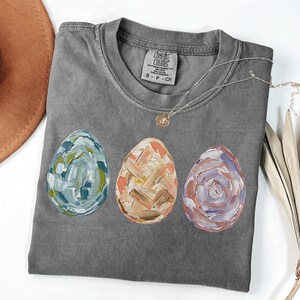 Peut inclure: Un t-shirt gris Comfort Colors avec trois &oelig;ufs de P&acirc;ques peints en bleu, orange et violet. Les &oelig;ufs pr&eacute;sentent un motif textur&eacute; au pinceau. Un collier argent&eacute; avec un petit pendentif est drap&eacute; sur le t-shirt.