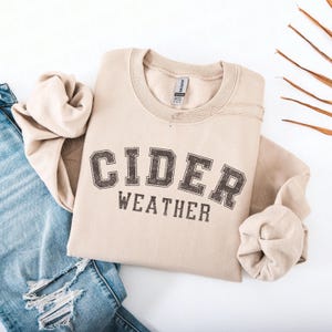 K&ouml;nnte beinhalten: Beiges Sweatshirt mit dem Aufdruck "CIDER WEATHER" in verblichener, dunkelbrauner Schrift. Das Sweatshirt liegt gefaltet auf einer wei&szlig;en Oberfl&auml;che neben einer Jeans.