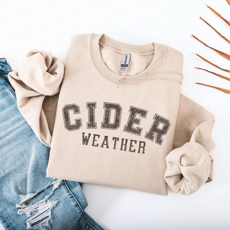 K&ouml;nnte beinhalten: Beiges Sweatshirt mit dem Aufdruck "CIDER WEATHER" in verblichener, dunkelbrauner Schrift. Das Sweatshirt liegt gefaltet auf einer wei&szlig;en Oberfl&auml;che neben einer Jeans.