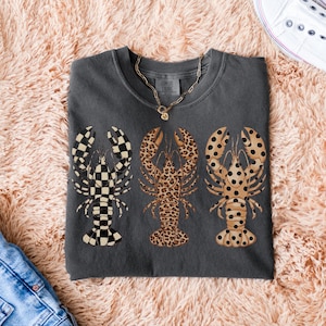 May include: Dark gray t-shirt featuring three lobster designs. One lobster has a black and white checkered pattern, the second has a leopard print, and the third has a tan and black polka dot pattern. A gold necklace is visible.