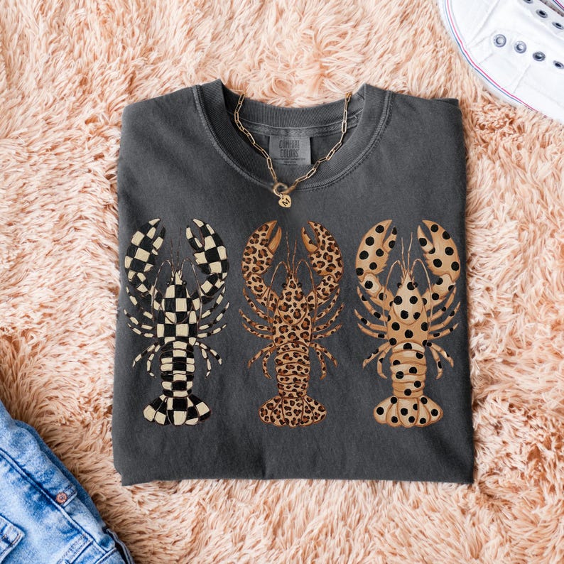 May include: Dark gray t-shirt featuring three lobster designs. One lobster has a black and white checkered pattern, the second has a leopard print, and the third has a tan and black polka dot pattern. A gold necklace is visible.