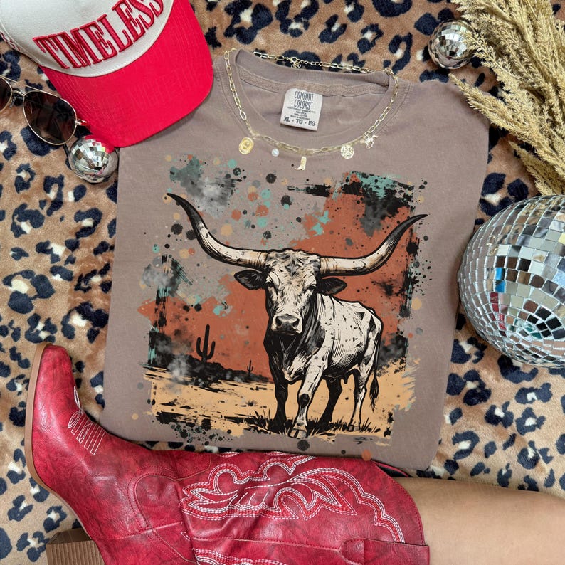 May include: A taupe t-shirt featuring a graphic of a longhorn steer with a desert landscape. The shirt is styled with a red baseball cap, sunglasses, a gold necklace, and red cowboy boots. The cap reads "Timeless".